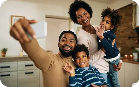 A happy family of four taking a selfie together in their kitchen, smiling and waving at the camera.