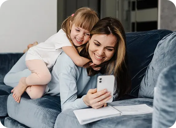 A smiling mother sitting on a couch with her young daughter hugging her from behind while they look at a smartphone together.